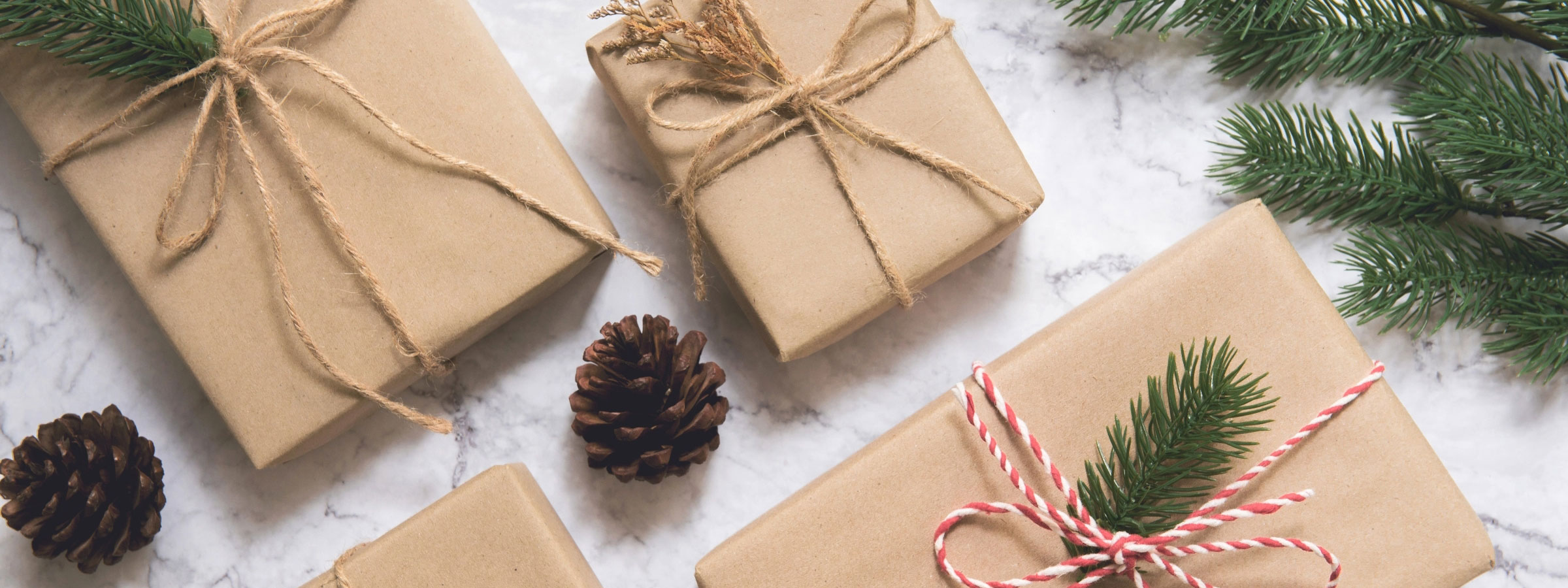 Three packages wrapped in brown paper with twine bows, with two pinecones and evergreen leaves.